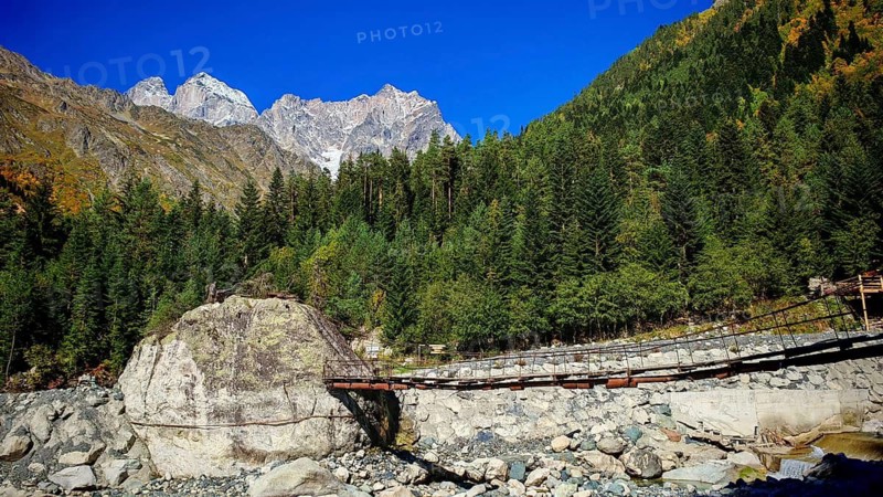 Suspension bridge over a rocky river surrounded by wooded mountain slopes and clear blue sky, hiking to Chalaadi Glacier, Mestia, Svaneti, Georgia