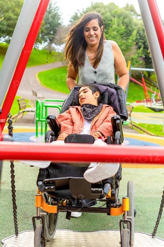A mother joyfully pushes her child with cerebral palsy in a wheelchair on a swing at an accessible park, surrounded by green trees. The image captures the essence of inclusion and outdoor fun