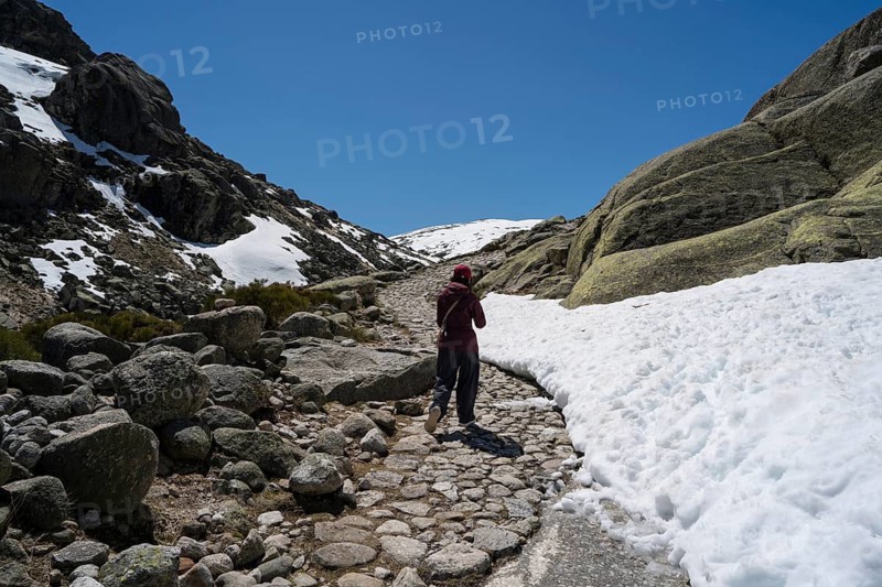 An Asian woman journeys on a snow-kissed path in Sierra de Gredos, Spain, surrounded by rugged mountains and clear blue skies, embracing adventure and nature