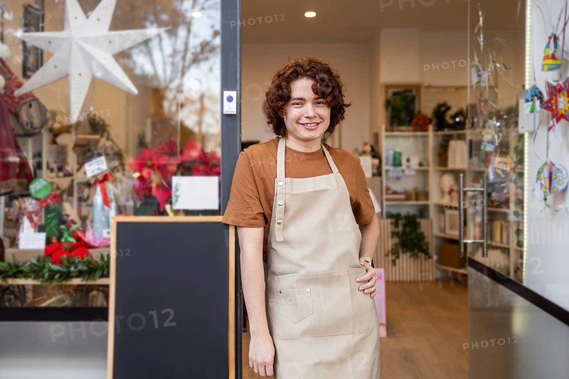 A cheerful shopkeeper in an apron stands at the entrance of their decorated store, inviting customers inside The scene conveys hospitality and holiday charm