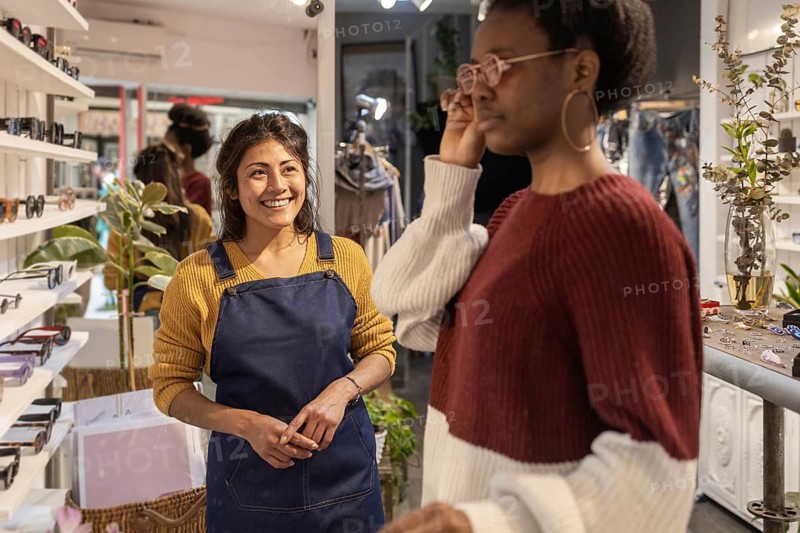A smiling store assistant in a modern sunglasses shop assists a customer trying on glasses. The boutique setting features stylish eyewear displays and a welcoming atmosphere