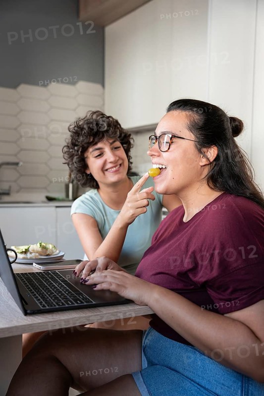 A joyful lesbian couple in a modern kitchen, sharing a light snack while one partner works on a laptop. The setting is cozy, highlighting their bond and domestic bliss