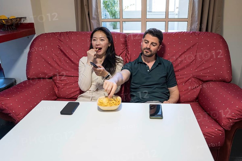 A diverse couple sits comfortably on a red sofa, enjoying snacks with a remote control as they relax at home, creating a cozy ambiance in their living space