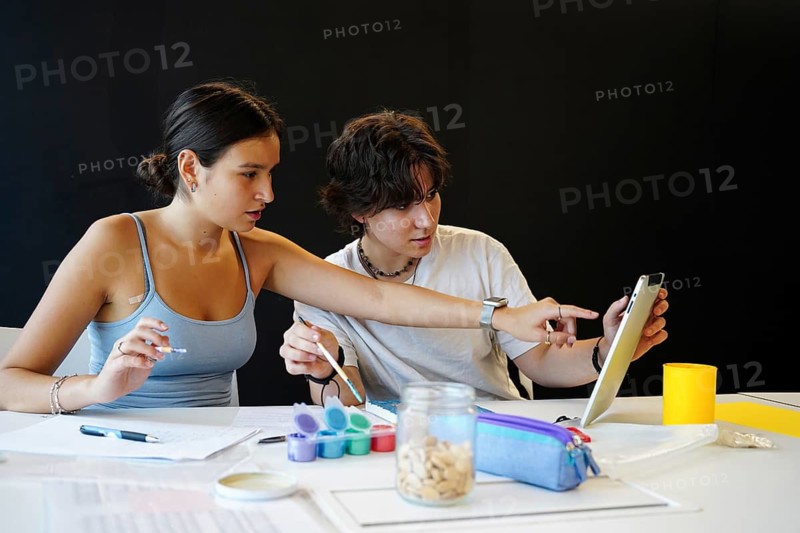 Female and non-binary teenager studying together, focused on a digital tablet and surrounded by artistic supplies. They engage in a collaborative and educational activity in a classroom setting