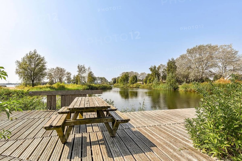 A tranquil scene featuring a wooden picnic table on a deck overlooking a serene lake surrounded by lush greenery and trees under a clear blue sky. Ideal for relaxation