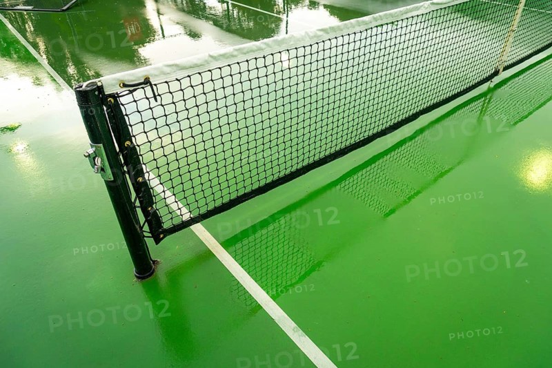 Pickleball court net standing on a vibrant green court surface wet from recent rain, reflecting surrounding light and creating dynamic shadows on the ground
