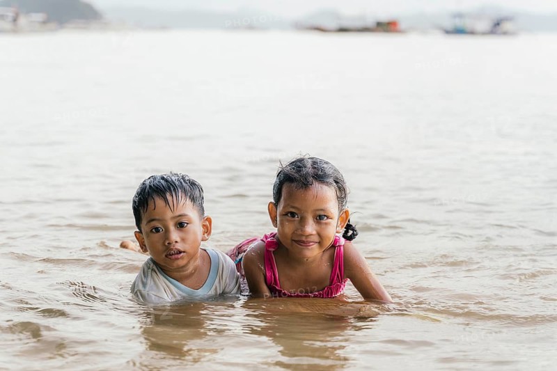 Two children joyfully play in the shallow water at a beach in the Philippines, looking at camera, embracing the fun of a sunny day. They smile, enjoying the warm ocean waves
