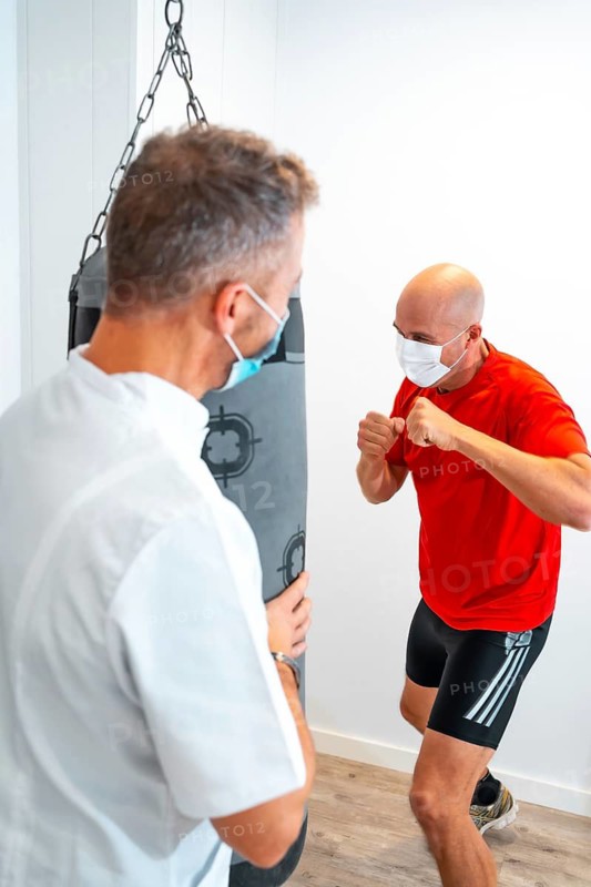 Physiotherapist with mask together with the patient on the punching bag. Physiotherapy with protective measures for the Coronavirus pandemic, COVID-19. Osteopathy, sports quiromassage