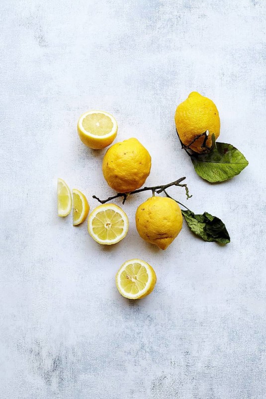 Top view of bright yellow lemons, some whole and some sliced, artistically arranged on a textured grey background. The fresh, vibrant lemons with green leaves evoke a natural, fresh feel, perfect for culinary and lifestyle themes