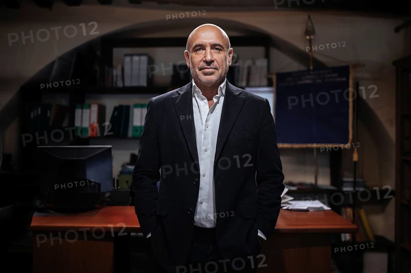 A confident businessman stands in a professional office setting. The background features shelves with files and a desk, creating an atmosphere of authority and focus