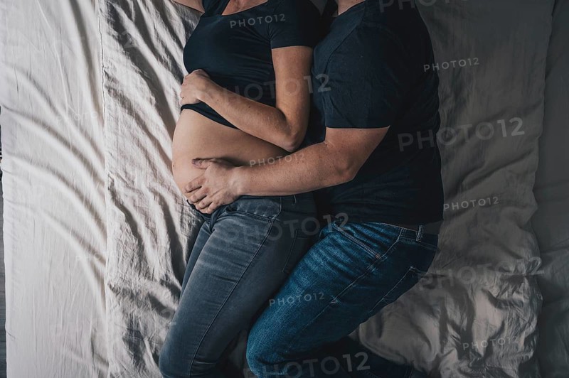 A couple is lying in bed, with the man embracing the pregnant woman and placing his hand on the baby's belly