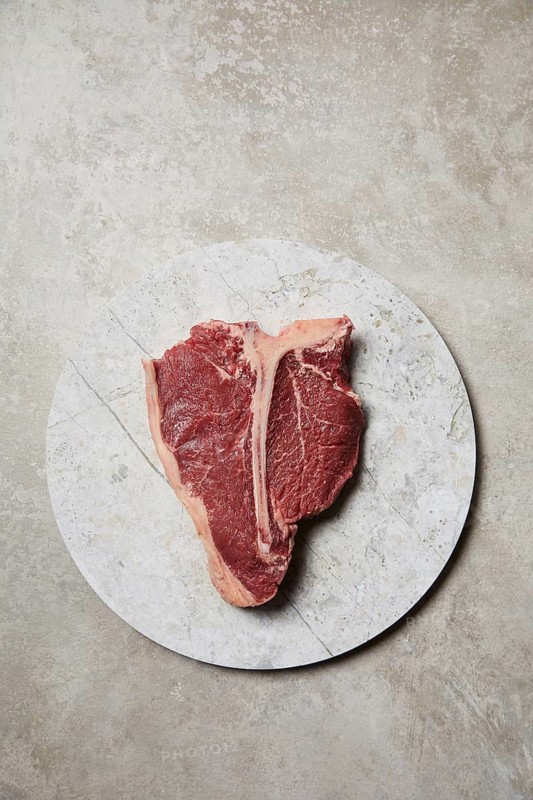 Freshly cut t-bone steak presented on a round marble plate, showcasing its juicy marbling and rich color, placed against a rustic textured backdrop for an inviting culinary display