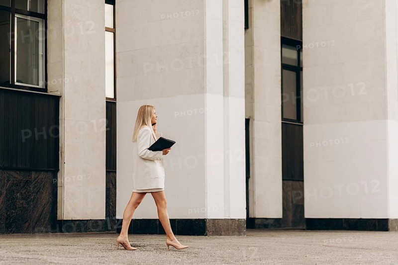 A business professional in formal attire confidently walks holding a folder, set against a modern office building backdrop, embodying corporate elegance and determination