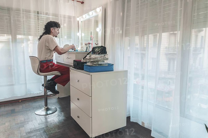 Young make-up artist applying cosmetics in front of a mirror inside a beauty salon, sitting comfortably on a stool beside a drawer unit, surrounded by tools and bright light