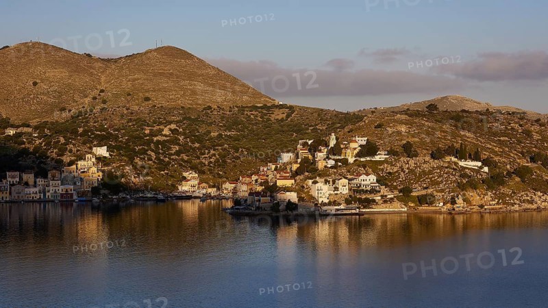 Coastal landscape with houses on the hill, illuminated by warm sunlight, calm water in the foreground, Symi Town, Symi, Dodecanese, Greek Islands, Greece