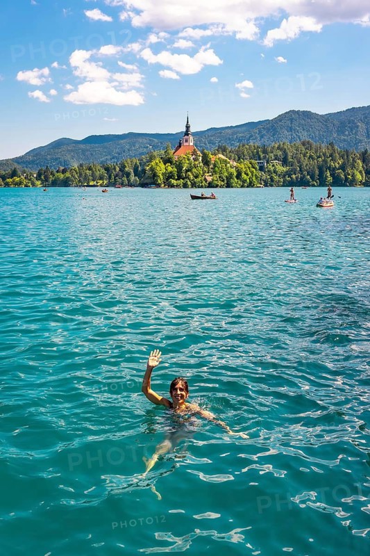 Tourist swimming in the turquoise waters of lake bled, waving at the camera with the assumption of mary church on the island behind