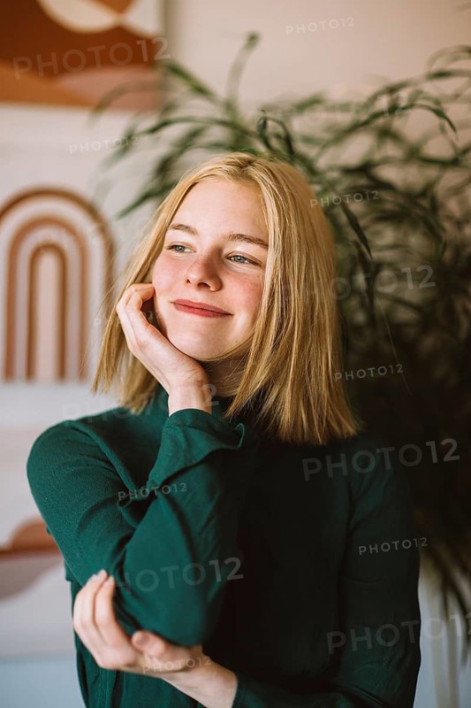 A teenager with blonde hair smiles thoughtfully, touching her face. She's wearing a green top in a cozy setting, conveying a sense of relaxation and happiness