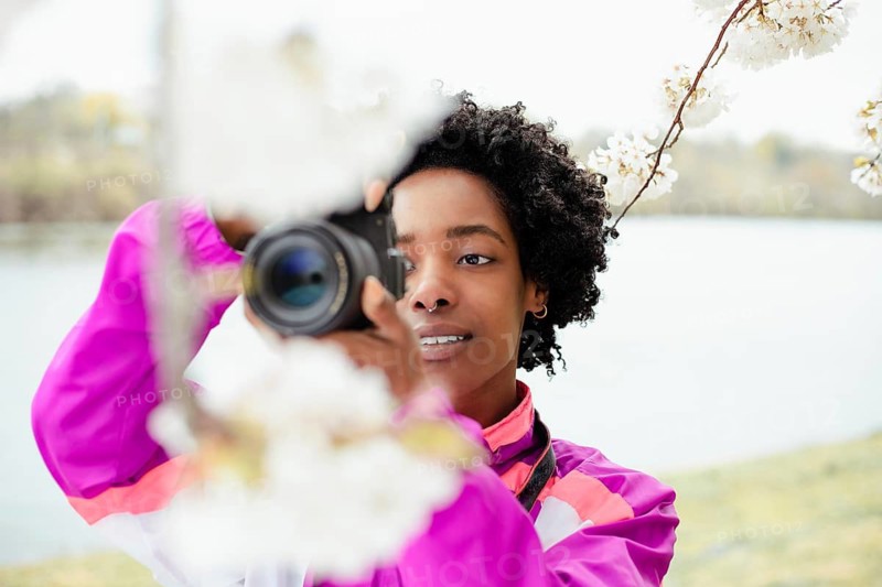A talented African American woman photographer skillfully captures the beauty of cherry blossoms with her camera. She stands outdoors, wearing a colorful jacket
