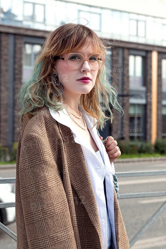 Stylish woman with wavy hair, wearing glasses and a blazer, poses confidently in an urban environment Her casual yet chic look highlights her individuality and fashion sense