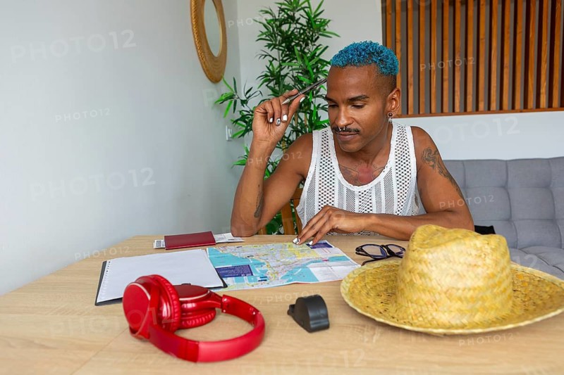An LGBTQIA+ man with vibrant blue hair is preparing for a trip. He is seated at a table with a map, hat, and headphones, representing travel and adventure readiness