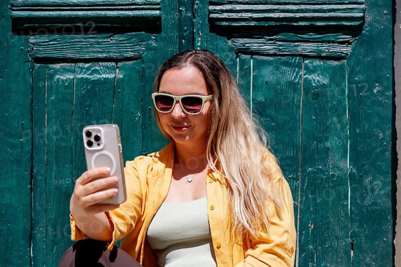 A smiling woman tourist wearing sunglasses and a yellow shirt takes a selfie on a colorful street, capturing her travel memories against a stylish, rustic backdrop