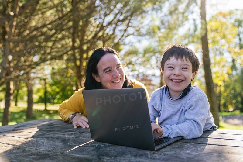 A smiling boy with Down syndrome and his mother enjoying a learning session outdoors with a laptop. Sunlight filters through trees, creating a warm and inviting atmosphere