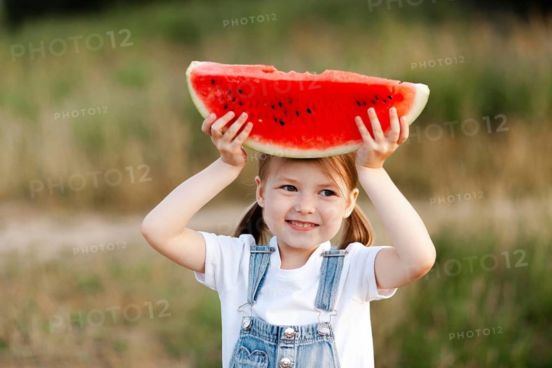 A joyful child holds a slice of watermelon overhead in a sunny summer field. Capturing the essence of carefree childhood, this image embodies the simple pleasures of warm seasons