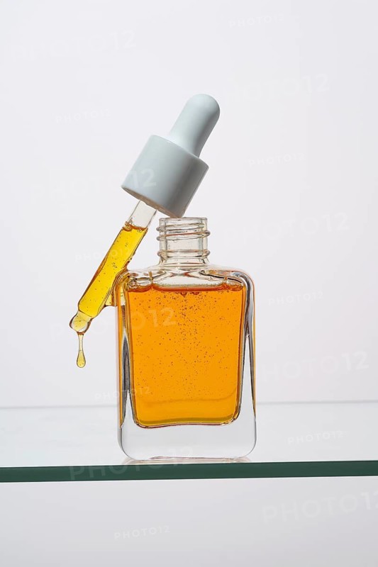 A clear bottle of vitamin C serum with a dropper is positioned on a glass surface The orange serum is delicately captured dripping from the pipette, highlighting its smooth texture