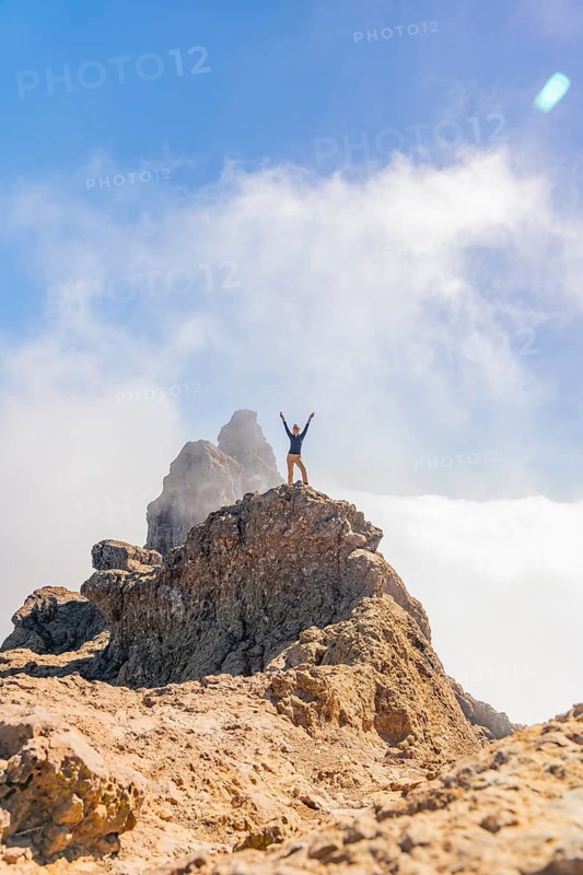 Distant excited woman with arms raised in victory on the rocky summit of Pico de las Nieves, Gran Canaria, surrounded by clouds and blue sky
