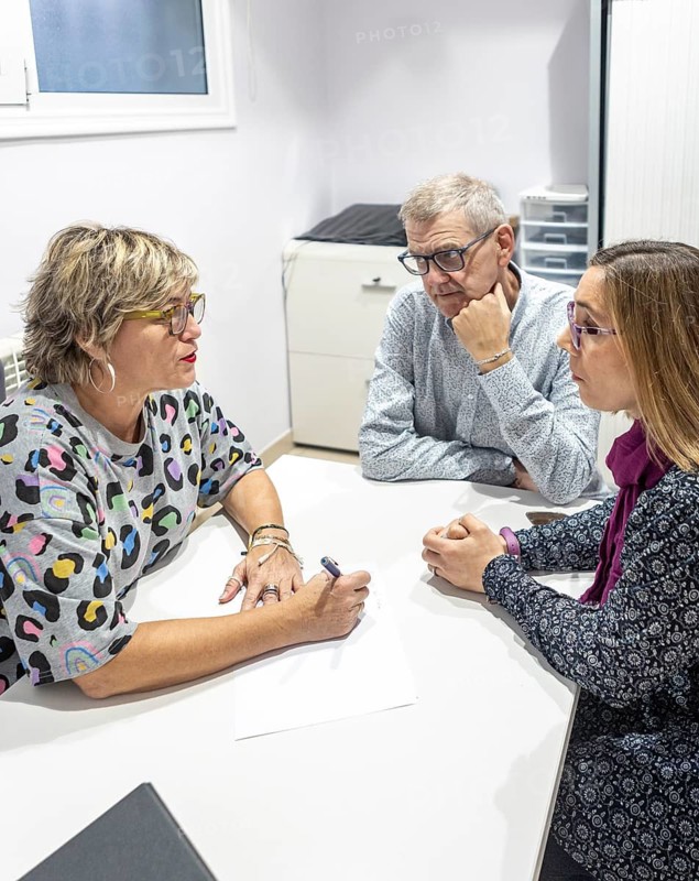 A woman and a couple with intellectual disabilities sit in an supervision meeting, engaged in conversation. A woman takes notes while the couple listen intently