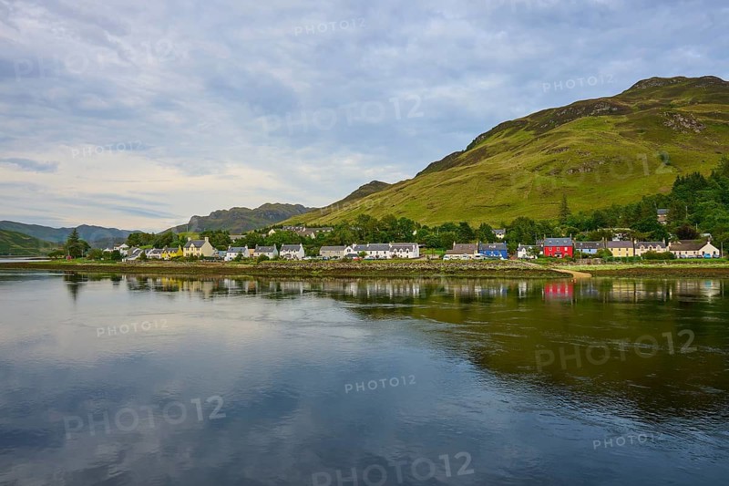 A calm lake with mountains and houses on the shore surrounded by green trees under a cloudy sky, Dornie, Kyle of Lochalsh, Scotland, United Kingdom, Great Britain