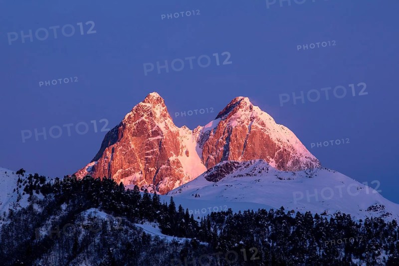 Snow-capped peaks glow with warm sunset hues against a vibrant blue sky in Georgia A dense forest covers the lower slopes, creating a breathtaking natural landscape