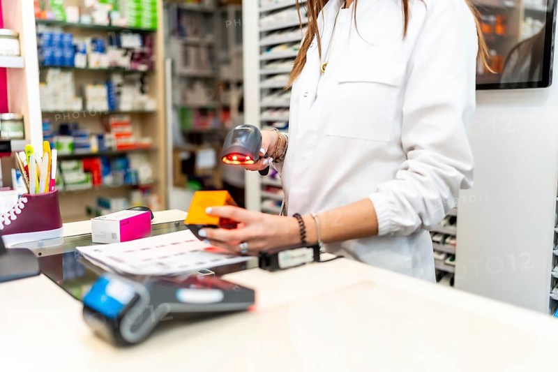 Pharmacist scanning medication packaging with a barcode reader at the pharmacy counter, preparing to complete a transaction for a customer with shelves of medicines in the background