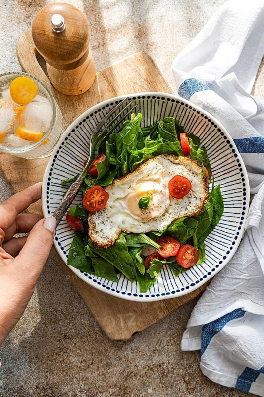 Top view of a healthy lunch featuring fresh spinach leaves, cherry tomatoes, and a fried egg, accompanied by a glass of chilled mineral water with ice and kumquat slices