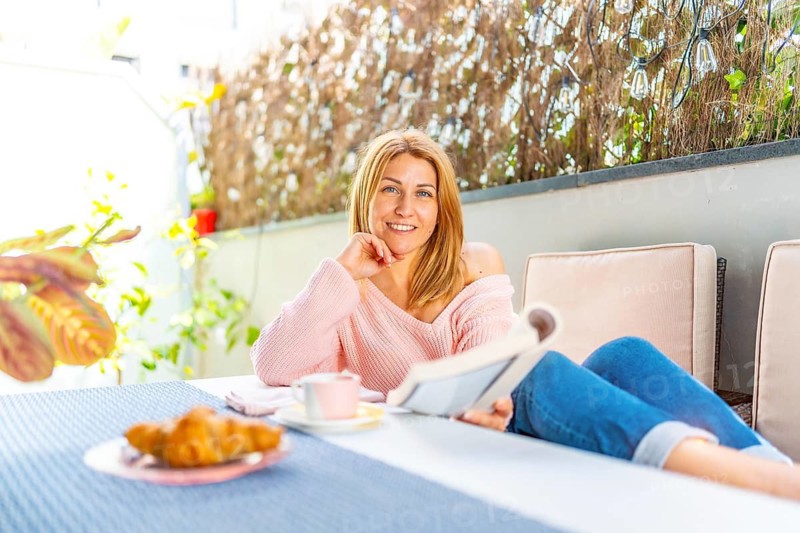 Adult caucasian blonde woman smiling at camera lying on a sofa in the outdoor yard enjoying breakfast and reading a book