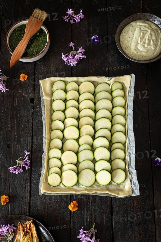 A beautifully arranged zucchini tart, complemented by vibrant edible flowers and bowls of green herb sauce and creamy dip, set on a dark, rustic wooden table