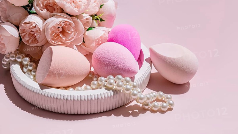 Various makeup egg sponges, a beauty blender, arranged amidst faux pearls and pink roses on a light pink background