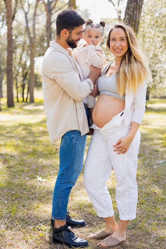 A happy pregnant woman stands in a sunny outdoor setting with her family. Her partner and young child share joyous moments, surrounded by nature and trees in the background