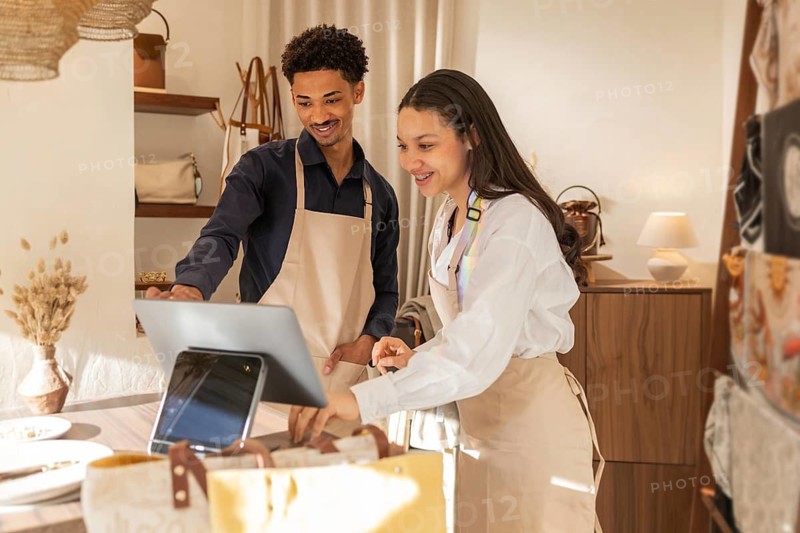 Two cheerful employees in a bag store using a tablet for checkout. They're standing behind the counter, surrounded by various bags and accessories on display