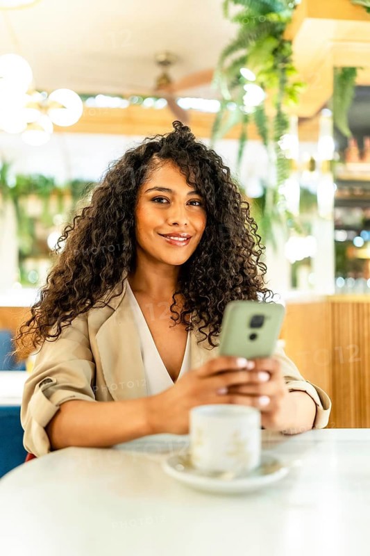 Vertical portrait of a chic beauty latin woman using phone sitting on a cafeteria drinking hot coffee