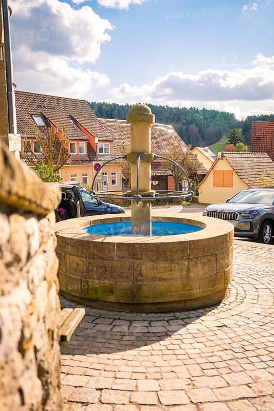 A round stone fountain in a village street surrounded by parked cars and houses under cloudy sky, Deufringen, Aidlingen, Böblingen district, Germany