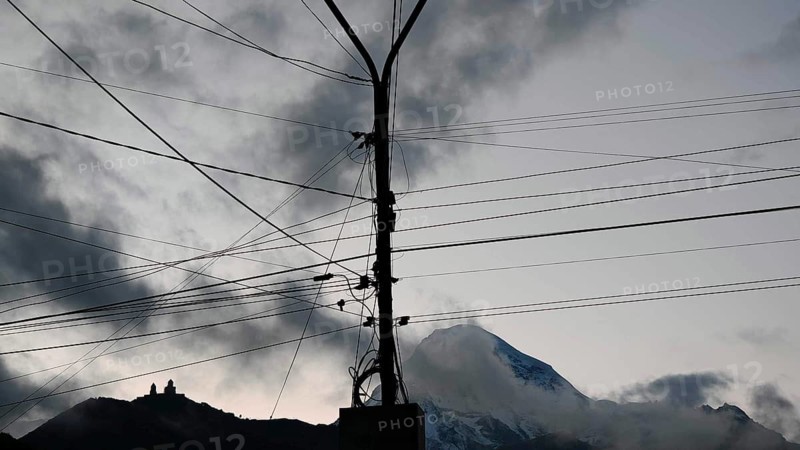 A power pole with cables in front of a snowy mountain, Kazbek with Gergeti church under dramatic clouds, Kazbegi, Stepantsminda, Georgia