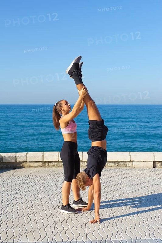 Fit woman helps man maintain a handstand near a scenic seaside promenade. Both wear sportswear, and the blue ocean creates a calm yet energetic background for fitness practice