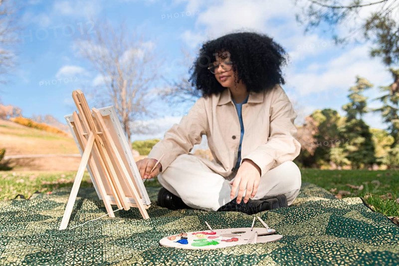 A young black artist sits on a patterned blanket in a park, painting on a canvas set on an easel. A vibrant paint palette rests nearby, capturing the essence of outdoor creativity