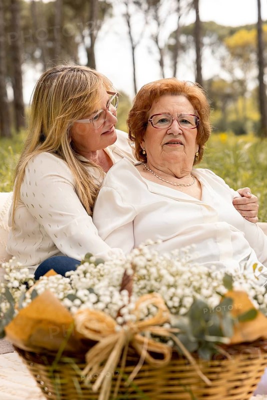 An adult daughter lovingly embraces her elderly mother as they spend quality time together outdoors, surrounded by nature and a basket of flowers, fostering a moment of connection