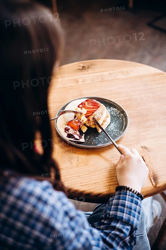 A woman in a plaid shirt uses utensils to enjoy a japanese pancakes adorned with sliced strawberries and syrup, served on a round plate, creating a cozy dining experience