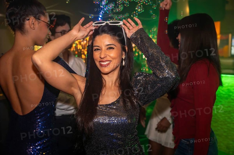 Young woman smiling confidently with white sunglasses on her head, enjoying a vibrant night out with friends amid sparkling lights and floating bubbles, celebrating youthful joy