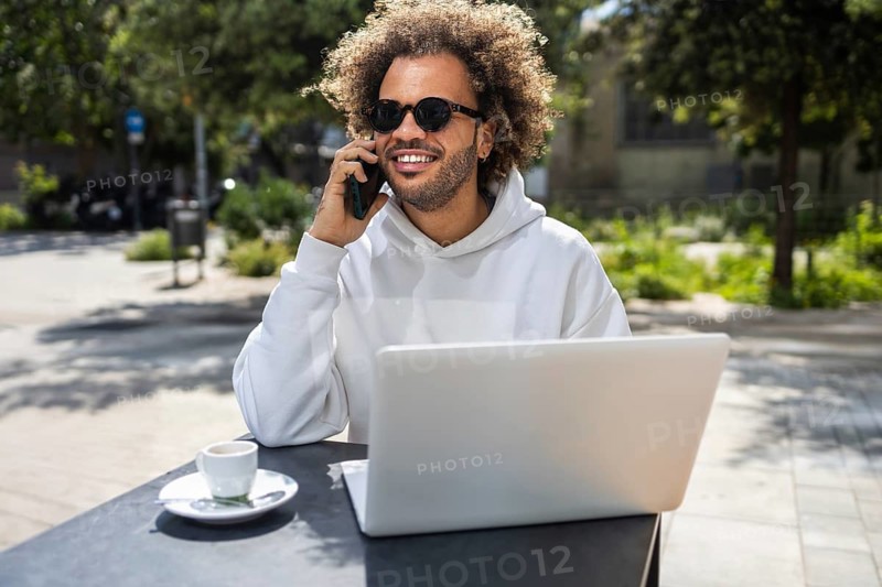 A cheerful man with curly hair smiling while talking on the phone at an outdoor cafe, sitting by his laptop with a cup of coffee. The scene reflects a relaxed atmosphere