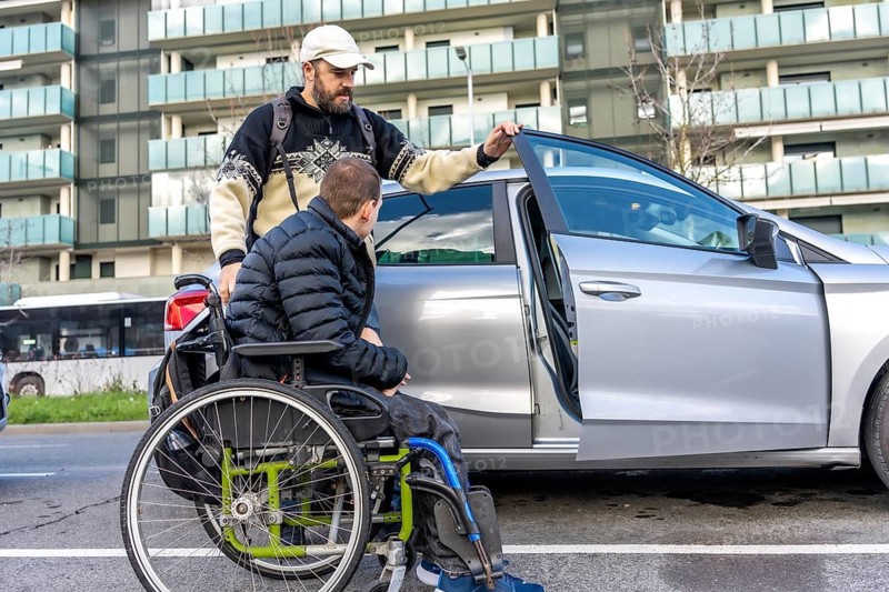 Caregiver assisting a young disabled man in a wheelchair while entering a car parked on a city street