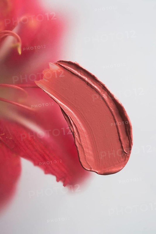 A vibrant smear of red lipstick is captured in a close-up, set against a softly blurred red flower against white background, highlighting the texture and color